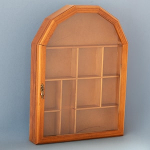 May include: A wooden display cabinet with a clear glass front and multiple shelves. The cabinet has a light brown finish and a decorative brass-coloured latch. The cabinet is designed to showcase small collectibles or decorative items.