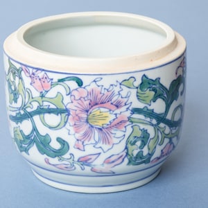 May include: A white ceramic planter with a floral design. The planter features a pink and yellow flower with green leaves and blue accents. The rim is a light beige colour, and the base has blue stripes.