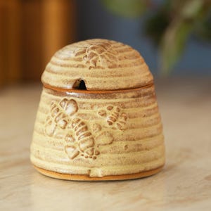May include: A ceramic honey pot with a lid, featuring a beehive design. The pot is tan with brown accents and embossed bee motifs. The lid has a small opening for a spoon. The pot is approximately 10 cm tall.