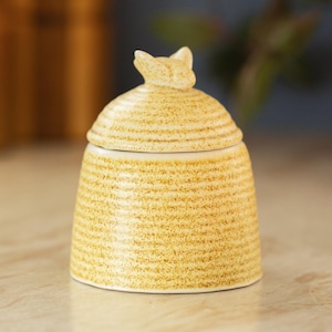 May include: A ceramic honey pot with a lid, featuring a textured, yellow and brown speckled design. The lid is topped with a small, decorative bee. The pot has a round base and a cylindrical body, ideal for kitchen decor.