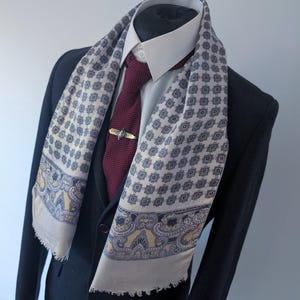 Puede incluir: Un conjunto formal con una blazer azul marino, una camisa blanca y una corbata burdeos. Una bufanda estampada en crema, azul y amarillo está drapeada alrededor del cuello. Un clip de corbata dorado con un elemento decorativo.