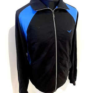 Puede incluir: Chaqueta negra con cremallera y mangas de color azul a contraste. La chaqueta tiene un pequeño logotipo en el pecho izquierdo.