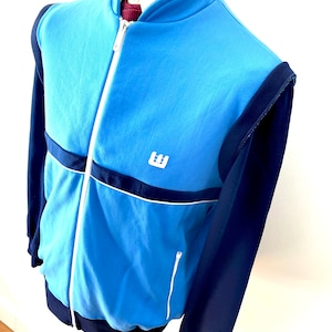 Puede incluir: Una chaqueta azul y azul marino con cremallera, con ribetes blancos y un logotipo blanco en el pecho. La chaqueta tiene un cuello alto y mangas largas.