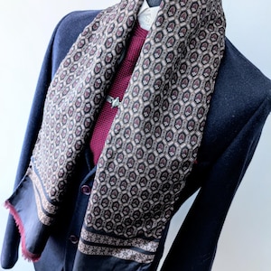 May include: A long scarf featuring a repeating geometric hexagonal pattern in shades of black, white, and deep red. The scarf has a solid black border with a thin decorative band and a frayed, deep red edge, draped over a dark navy blazer and shirt.