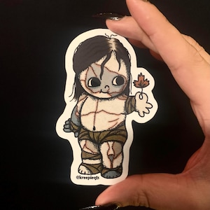 May include: A sticker featuring a cartoon-style zombie baby holding a brown leaf. The baby has stitched-up skin, dark hair, and is wearing wrapped bandages. The sticker has the text "@kreepieqts".