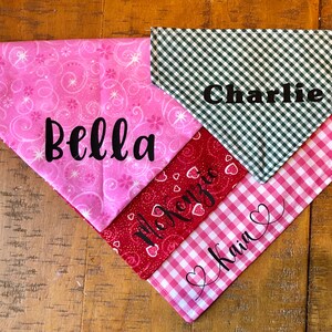 May include: Three dog bandanas with different patterns and names. The top bandana is green and white gingham with the name "Charlie" embroidered on it. The middle bandana is red with white hearts and the name "Kenzie" embroidered on it. The bottom bandana is pink and white gingham with the name "Kain" embroidered on it.