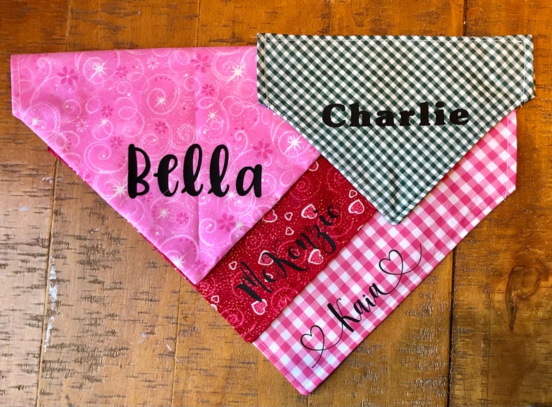May include: Three dog bandanas with different patterns and names. The top bandana is green and white gingham with the name "Charlie" embroidered on it. The middle bandana is red with white hearts and the name "Kenzie" embroidered on it. The bottom bandana is pink and white gingham with the name "Kain" embroidered on it.