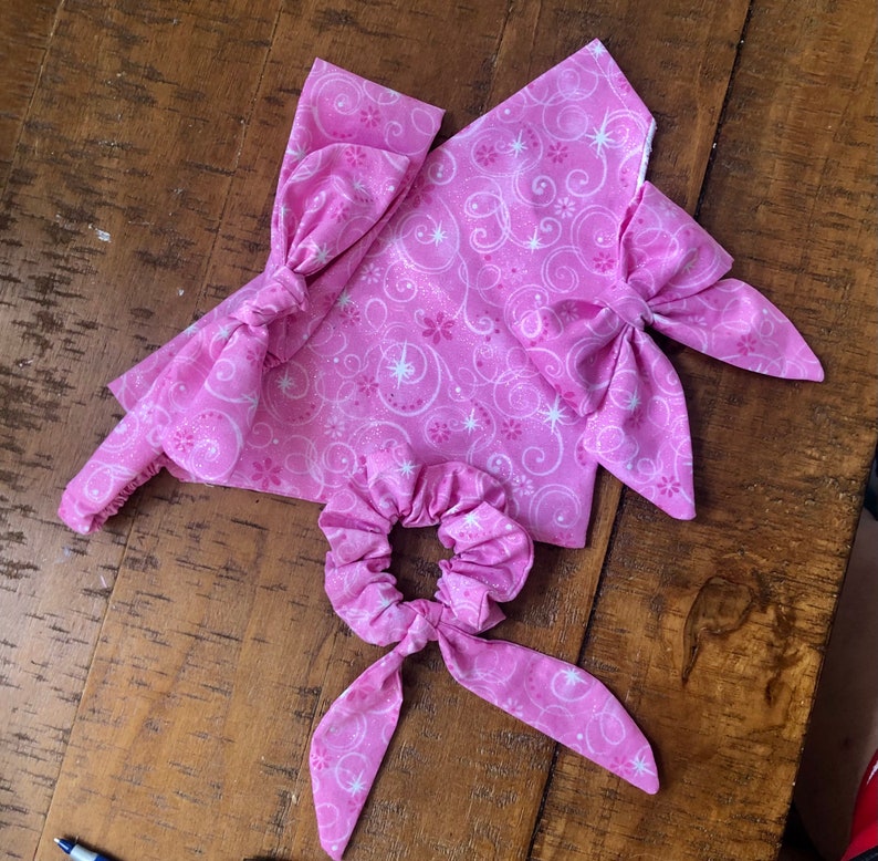 May include: Pink fabric hair scrunchie with a bow and a pink and white patterned design. The scrunchie is tied in a bow.