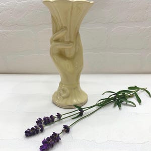 May include: A cream-colored ceramic vase shaped like a hand holding a flared opening. The vase is displayed with two sprigs of lavender with purple flowers and green stems.