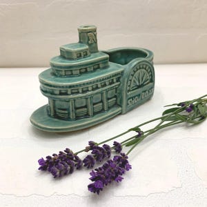 May include: A teal ceramic steamboat planter with the word "SHOWBOAT" on the side. The planter has a detailed design and is accompanied by sprigs of purple lavender.