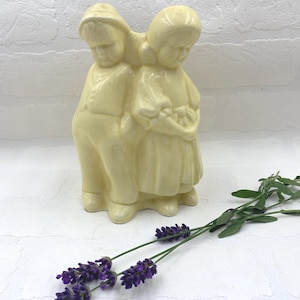 May include: A vintage ceramic figurine depicting two children, a boy and a girl, in a cream color. The girl is holding a basket. The figurine is next to sprigs of purple lavender.
