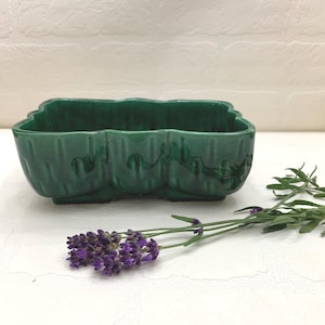 May include: A green ceramic planter with a scalloped edge. The planter has a three-section design and is decorated with embossed details. A sprig of purple lavender rests beside the planter.