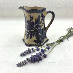 May include: A ceramic pitcher with a cream-colored base and dark blue floral patterns. The pitcher features a scalloped rim, a curved handle, and a flared base. Beside the pitcher lies a sprig of purple lavender with green stems.