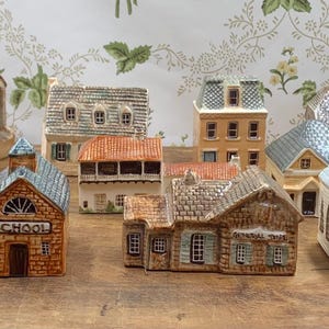 May include: A collection of miniature ceramic houses and buildings, including a school, church, and various homes. The buildings feature detailed architectural elements and are painted in a variety of colors, including brown, beige, and blue.