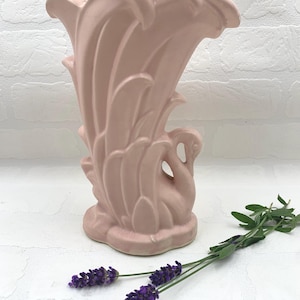 Pode incluir: Um vaso de cerâmica rosa com um desenho de cisne. O vaso tem um topo alargado e um cisne esculpido na frente. O vaso tem aproximadamente 20 cm de altura. Ramos de lavanda roxa estão ao lado do vaso.