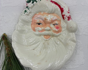 May include: A ceramic Santa Claus face wall plaque featuring a white beard, a red hat with green holly leaves, and pink cheeks. The Santa has a smiling expression with painted eyes and a glossy finish, showing signs of vintage paint wear.