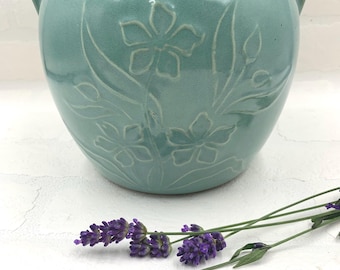 Puede incluir: Una maceta de cerámica color turquesa con diseños florales en relieve y dos asas pequeñas. La maceta es redonda y está sobre una superficie blanca, con ramitas de lavanda púrpura en primer plano. Un recipiente decorativo para plantas.