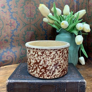 May include: A vintage stoneware crock with a speckled brown and cream pattern. The crock sits atop a dark, antique book. A vase of white tulips is in the background, against a patterned wallpaper.