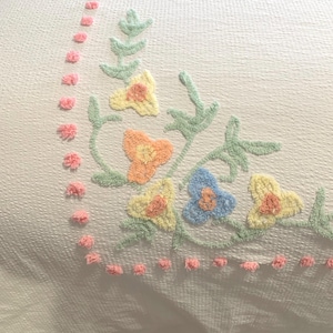 May include: A white crocheted bedspread with a floral design in pastel colours. The flowers are raised and have a fluffy texture. The bedspread has a pink pom-pom trim.