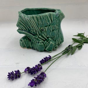 May include: A green ceramic planter shaped like a tree stump, with the word "STUV" carved into the side. The planter is decorated with small, embossed details. Two sprigs of lavender with purple flowers are in the foreground.