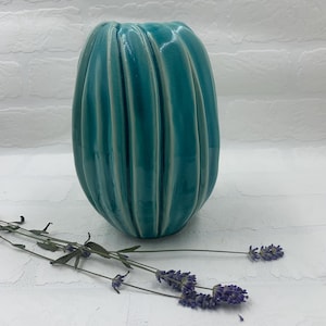 May include: A teal ceramic vase with vertical ribbed detailing. The vase has a glossy finish and an oval shape. Dried lavender sprigs are placed in front of the vase, adding a touch of natural color and texture.