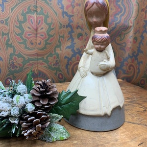 May include: Ceramic figurine of a woman holding a child. The woman is wearing a white dress with a gray base and a yellow head scarf. The child is wearing a white dress with a brown head. The figurine is on a wooden surface with a green and white pine sprig.