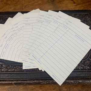 May include: A stack of vintage library card catalog cards fanned out on a dark, textured surface. Each card is white with blue lines and text fields for author, title, and date due. The cards are printed in the USA.