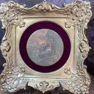 May include: Ornate gold-colored picture frame with a landscape painting. The frame has a detailed, scrollwork design and a burgundy velvet matting around the circular painting. The painting depicts a rural scene with a house, trees, and figures.