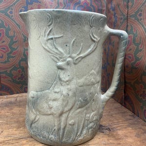 May include: A stoneware pitcher with a detailed relief of a stag in a naturalistic setting. The pitcher is a light beige color with blue-gray accents, featuring a handle and a spout. The design includes foliage and a mountain range.