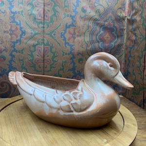 May include: A ceramic duck planter in shades of brown and white. The duck has a long, curved neck and a detailed wing design. The planter sits on a round wooden tray, with a patterned background.