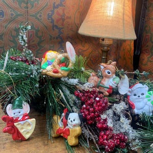 May include: A collection of Christmas ornaments, including a mouse, a dog, a reindeer, and a bear, arranged on a wooden surface with faux greenery and red berries. The ornaments are colorful and festive, with a vintage aesthetic.