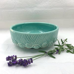 May include: A turquoise ceramic bowl with a textured design. The bowl has a rounded shape with a decorative pattern around the base. Sprigs of lavender are placed near the bowl, adding a touch of color and natural elements.