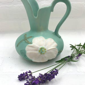 May include: A small, turquoise ceramic pitcher with a white flower design. The pitcher has a curved handle and a spout. A sprig of purple lavender is next to the pitcher.