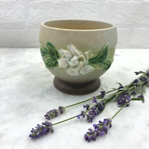 May include: A cream-coloured ceramic planter with a crackle glaze finish, featuring a raised relief of a white gardenia flower and green leaves around the centre. The bowl-shaped vessel sits on a dark brown circular base, accompanied by lavender.