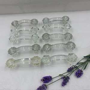 May include: A collection of eight clear glass cabinet pulls with a floral design on each end. The pulls are arranged in two rows of four, with a sprig of purple lavender in the lower right corner. The pulls are approximately 4 inches long.