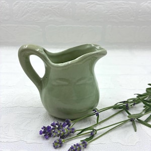 May include: A small, light green ceramic creamer pitcher with a curved handle and a pouring spout, accompanied by several sprigs of purple lavender flowers. The pitcher has a smooth, glossy finish and a rounded body shape with a wide opening.