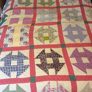 May include: A patchwork quilt featuring a grid of square blocks separated by red fabric sashing with small green corner squares. Each block contains a geometric pattern in various colours and prints, including floral, plaid, and solid fabrics.