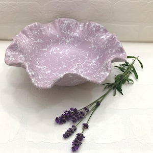 May include: A lavender-colored ceramic bowl with a ruffled edge and white splatter design. The bowl is displayed with a sprig of lavender and purple flowers, suggesting a decorative or serving piece.