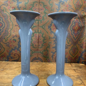 May include: A pair of light blue ceramic candle holders. Each has a flared top, a slender, curved stem, and a rounded base. The candle holders are displayed on a wooden surface.