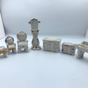 May include: A set of miniature white ceramic furniture, including a table, two chairs, a grandfather clock, two chests of drawers, and a piano. Each piece is decorated with colourful floral accents.