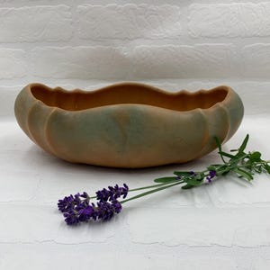 May include: A handcrafted, oval-shaped ceramic bowl with a scalloped rim. The bowl has a two-tone color scheme, with a muted green exterior and a warm brown interior. A sprig of lavender with purple flowers rests beside the bowl.