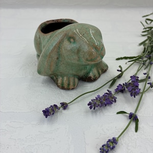 May include: A ceramic rabbit-shaped planter featuring a light green crackle glaze finish. The sculpture depicts a seated rabbit with long ears draped along the sides and a hollow opening on its back for small plants or decorative items.