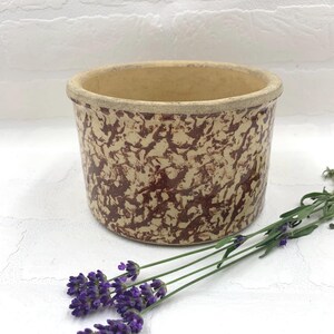 May include: A round, beige stoneware crock with a speckled maroon pattern. The rim is a lighter beige color. A sprig of purple lavender is placed next to the crock. The crock is likely used for storage or decoration.