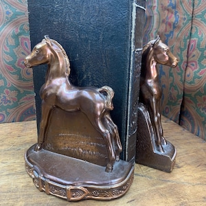 May include: A pair of bronze-colored horse bookends. Each bookend features a detailed horse sculpture on a decorative base. The bookends are positioned on a wooden surface, with a dark book between them. The background has a patterned fabric.