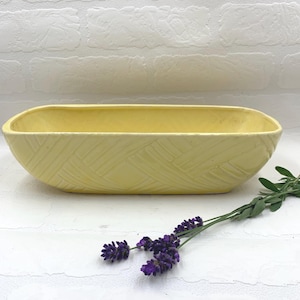 May include: A pale yellow rectangular ceramic planter with a textured woven pattern. The planter is empty and sits on a white surface. A sprig of purple lavender is placed near the planter.