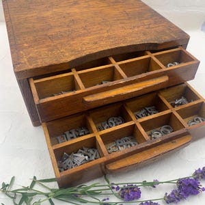 May include: A vintage wooden printer's drawer with two open drawers filled with metal letterpress type. The wood has a warm, aged appearance. A sprig of lavender with purple flowers rests in the foreground.