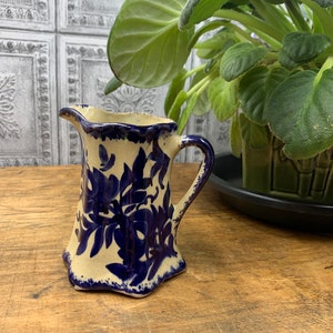 May include: A small, cream-coloured ceramic creamer with a blue floral design. The creamer has a scalloped rim and a handle.