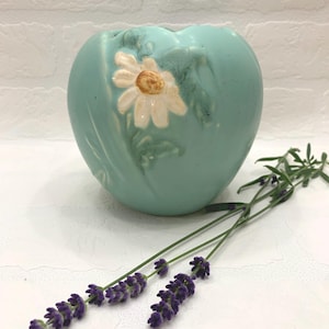 May include: A light teal ceramic vase with a daisy design. The vase has a rounded shape and a matte finish. A sprig of lavender with purple flowers rests in front of the vase.