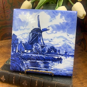 Peut inclure: Un carreau de céramique bleu et blanc représentant un moulin à vent hollandais avec un canal et des maisons au premier plan. Le carreau a un aspect rustique et vintage.