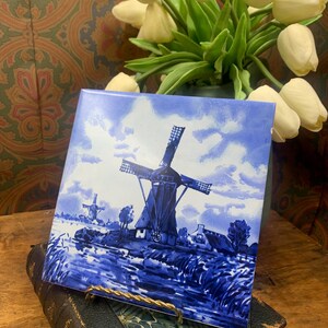 Peut inclure: Carreau en céramique bleu et blanc représentant un moulin à vent hollandais dans un paysage. Le carreau est présenté sur un support en métal doré.